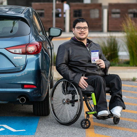 Young man in a wheelchair next to his car, holding up an ACROD permit.
